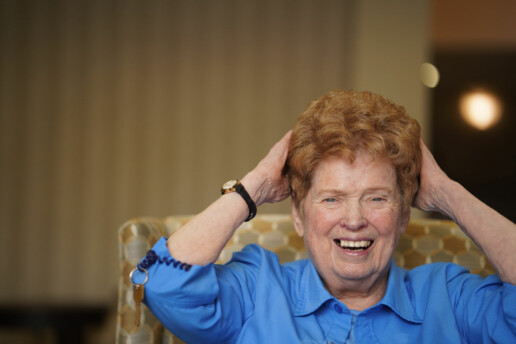 An smiling elderly woman in a blue blouse, touches her red hair while sitting in an upholstered lounge chair..
