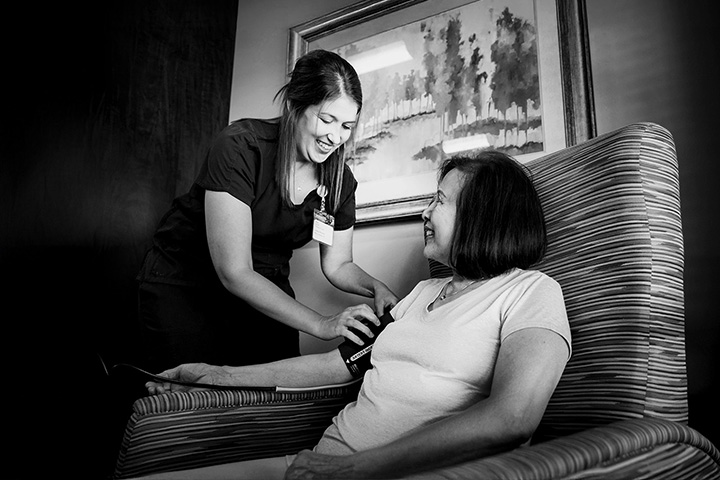 A healthcare worker, supported by corporate sponsors, checks the blood pressure of a seated woman. Both are smiling in a room with a framed painting on the wall, showcasing how community and sponsorships enhance well-being initiatives.