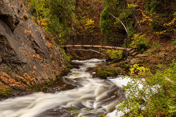 A wooden bridge spans over a rushing stream in a forest with autumn foliage and rocky cliffs.