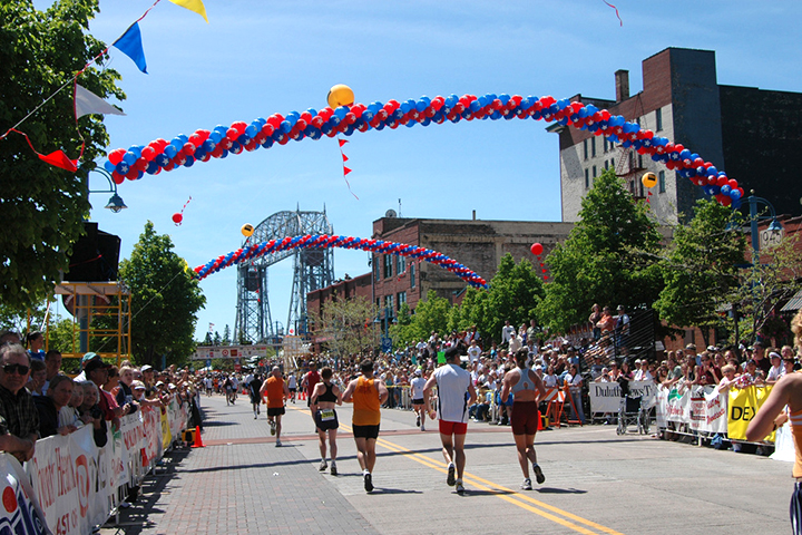 Runners in the Duluth Grandmas Marathon race under colorful balloon arches, with cheering crowds on a sunny day.