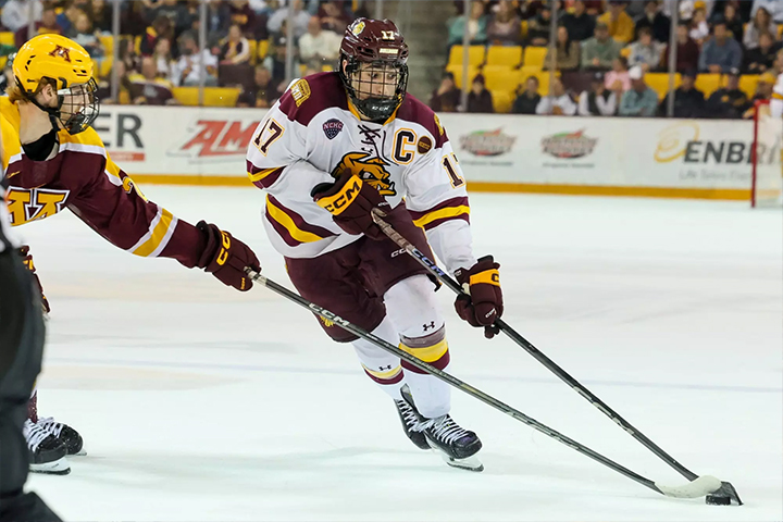 UMD Hockey player in maroon and white jersey, labeled C, skates with the puck on the ice, opponent behind.