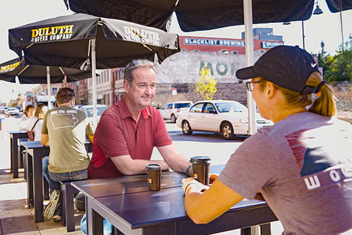 Two people sitting outdoors at a Duluth Coffee Company table, chatting and drinking coffee, with a street in the background.