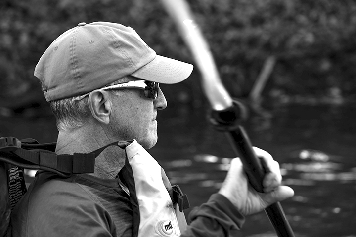 Man in a cap and sunglasses paddling in a kayak on a river, wearing a life jacket.