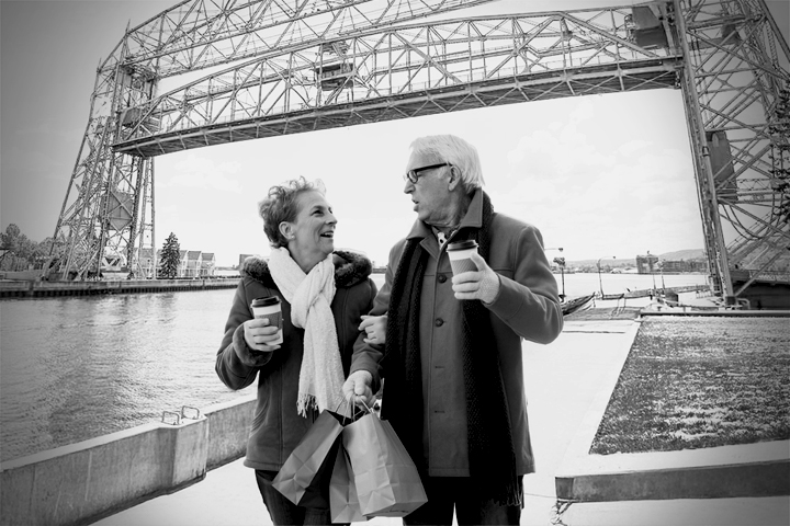 A smiling couple drinks coffee and carries shopping bags in front of the snowy Aerial Lift Bridge.