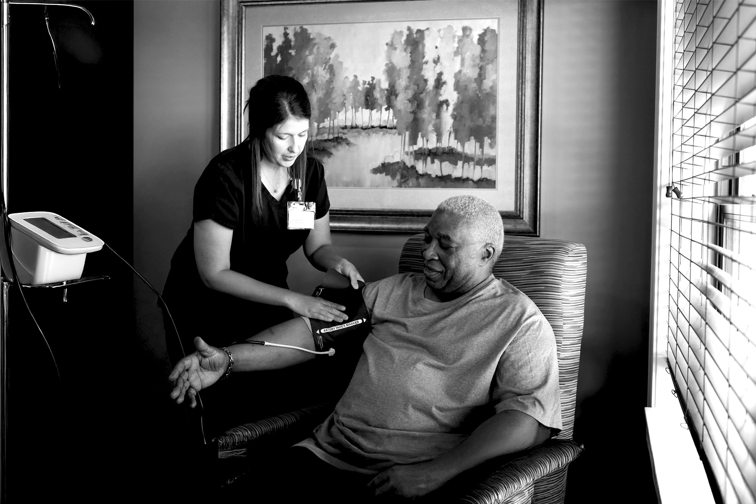 a caregiver checking the blood pressure of an older adult male sitting in an armchair near a window of a well lit room