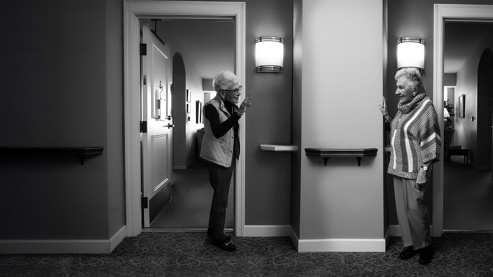 Two older women greet each other from separate neighboring doorways in a hallway, smiling and waving.