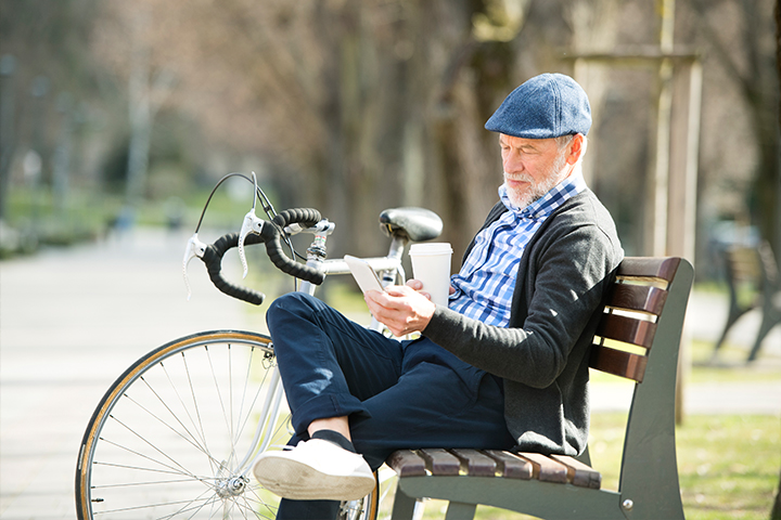Older man sitting on a park bench with a coffee, looking at his phone; bicycle leaned beside him.