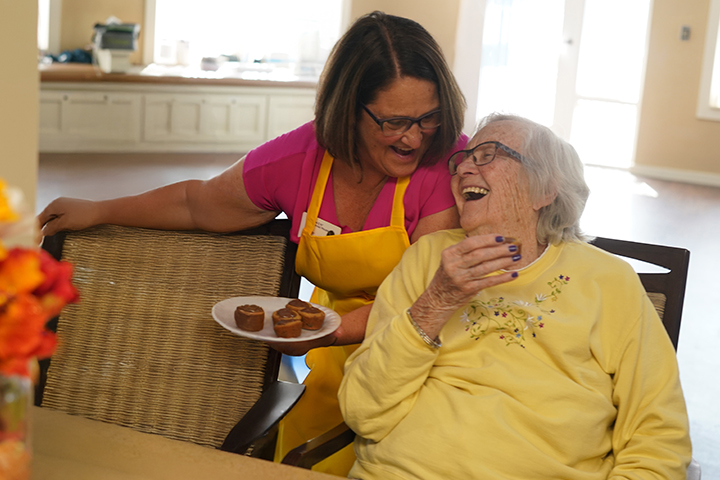 A caregiver serves muffins to a smiling elderly woman, both laughing and enjoying their time together.