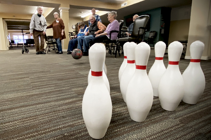 Elderly people play indoor bowling, aiming a ball at a set of white pins on a carpeted floor.