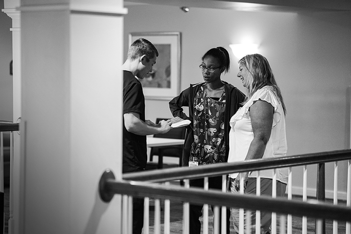 Three people stand together indoors, talking and smiling near a railing in a black-and-white photo.