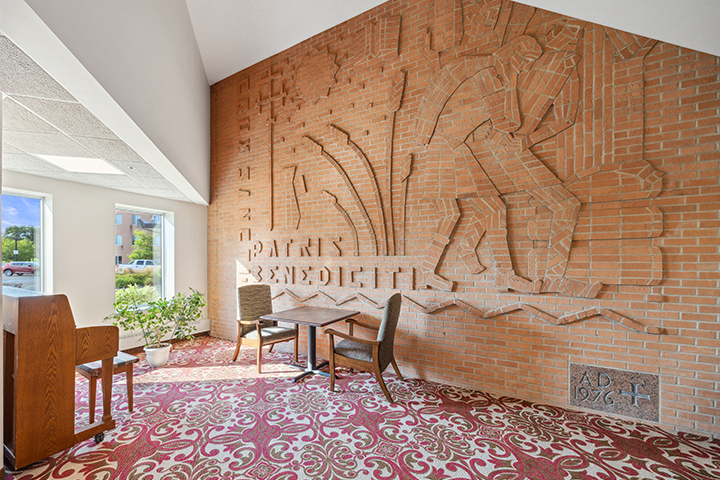 A brick wall with carved art, two chairs, a table, a piano, and patterned carpet in a bright room.