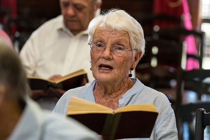 Elderly woman with white hair singing while holding a book, seated among others in a gathering.