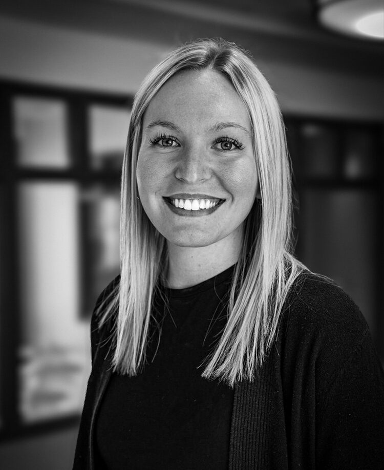Hailey Richter, asmiling woman with straight, blonde hair wearing a dark top, stands indoors with blurred background.