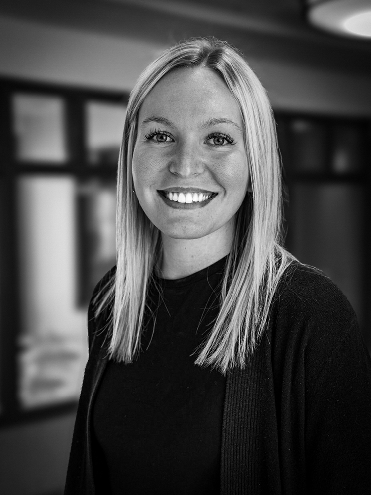 Hailey Richter, asmiling woman with straight, blonde hair wearing a dark top, stands indoors with blurred background.