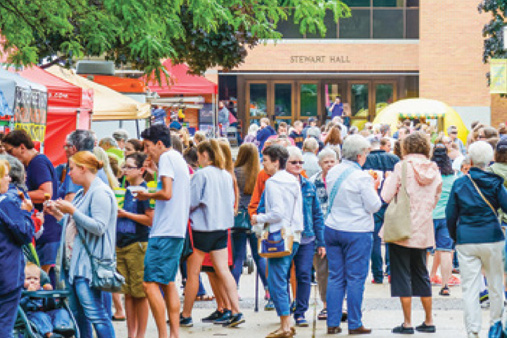 A crowd gathers outdoors at a lively art fair in front of a building labeled Stewart Hall.
