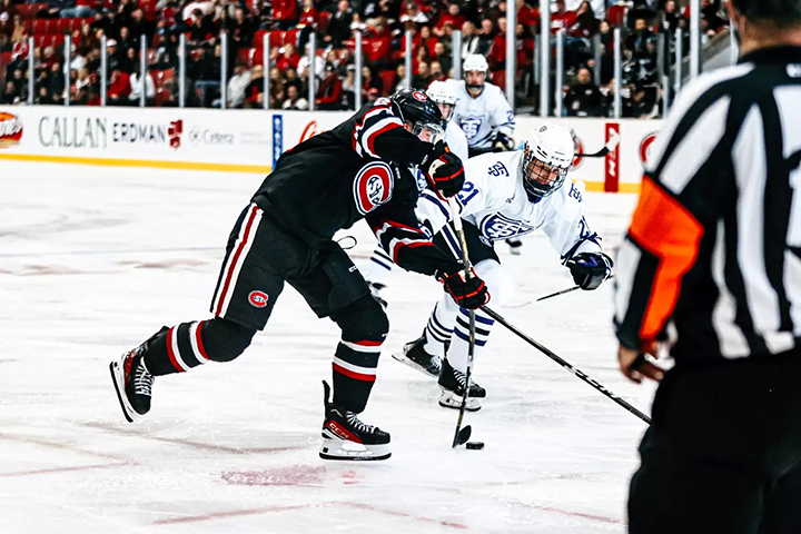 St. Cloud State University hockey players compete in a game against University of St. Thomas hockey players with a referee in the foreground and spectators in the background.