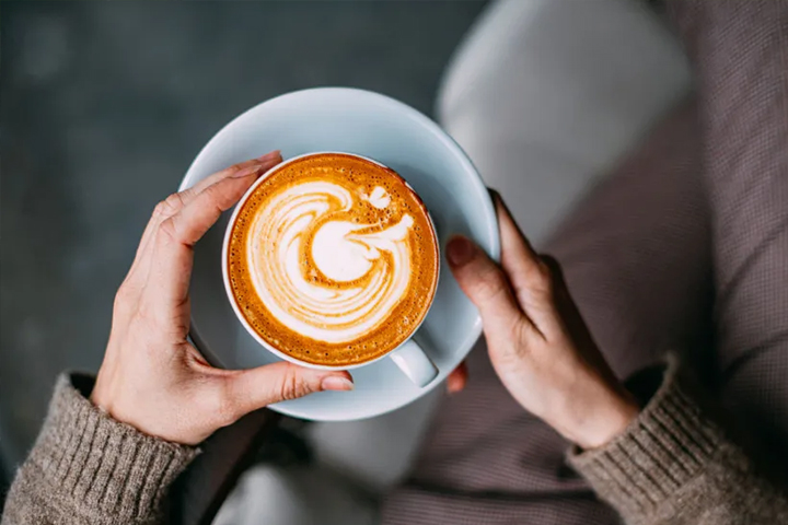 Person holding a cup of latte with swirled latte art on top, wearing a cozy brown sweater.