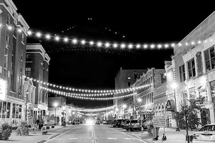 A city street at night with string lights overhead and buildings lining both sides, in black and white.