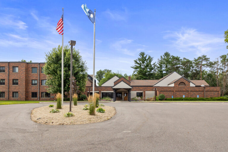 A brick building with a sloped roof marks the Benedict Village Entrance, surrounded by flags, a parking lot, and trees.