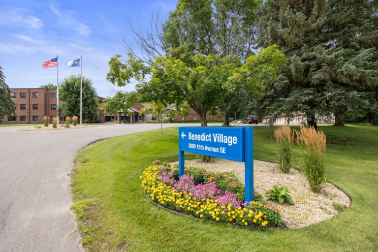 Blue sign for Benedict Village with green lawn, flowers, and two flagpoles; building in the background.