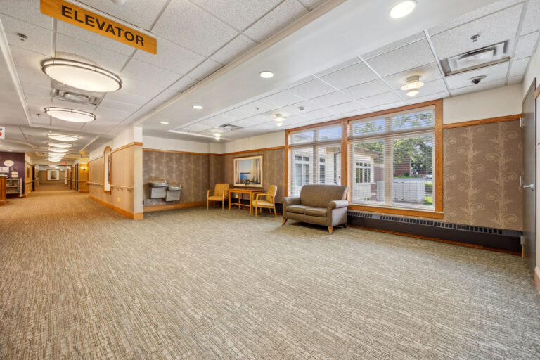 Long-term care facility lounge area includes spacious hallway with chairs, a table, and elevator sign,