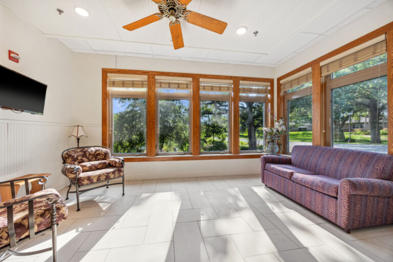 The long-term care sunroom is a sunlit living room with large windows, a sofa, and ceiling fan, overlooking a green lawn and trees.