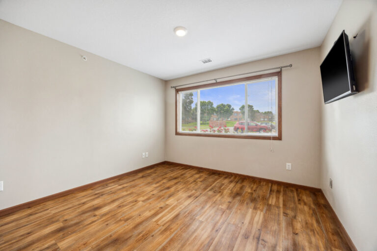 Benedict Homes studio with wooden floor, beige walls, wall-mounted TV, and large window showcasing trees.