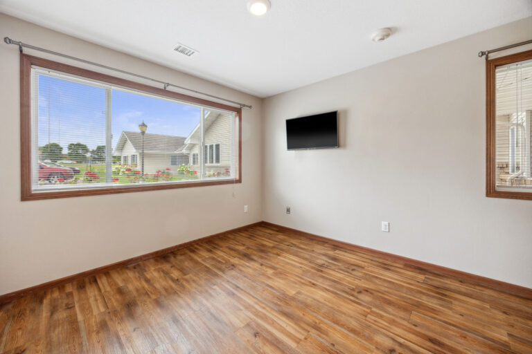 Benedict Homes studio with wooden floor, beige walls, wall-mounted TV, and large window showcasing trees.