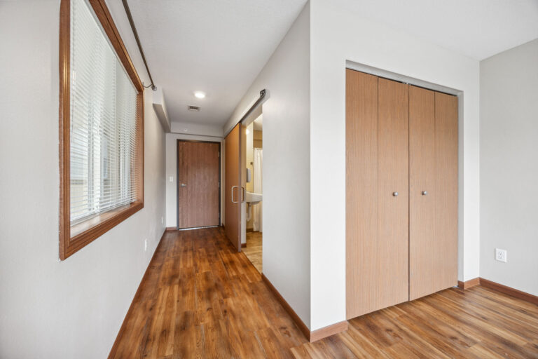 Interior hallway of Benedict Homes memory care apartment with wooden floors, a closet, and a bathroom visible at the end.