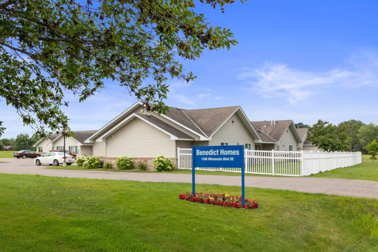 Memory Care building with a white fence, manicured lawn, and a blue sign reading Benedict Homes.
