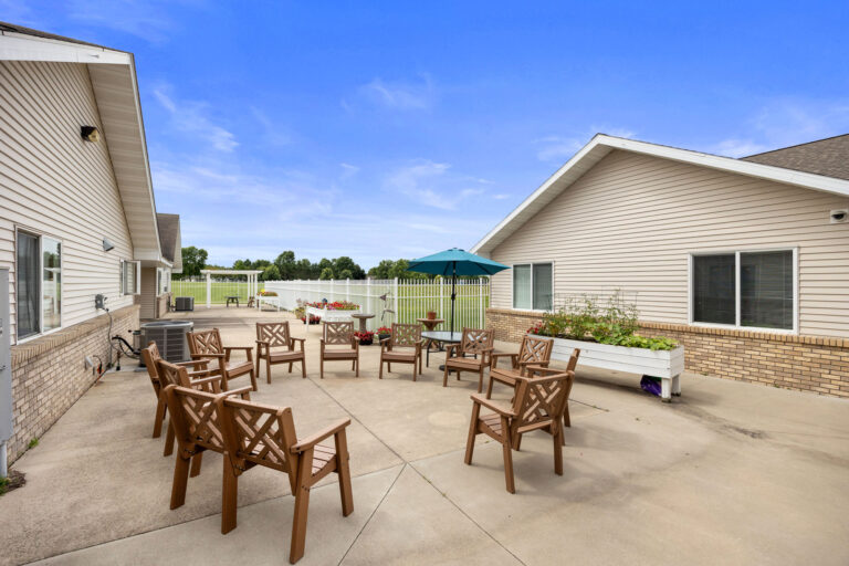 Chairs circle a green umbrella on the Benedict Homes patio, planters nearby, framed by beige buildings under a blue sky.