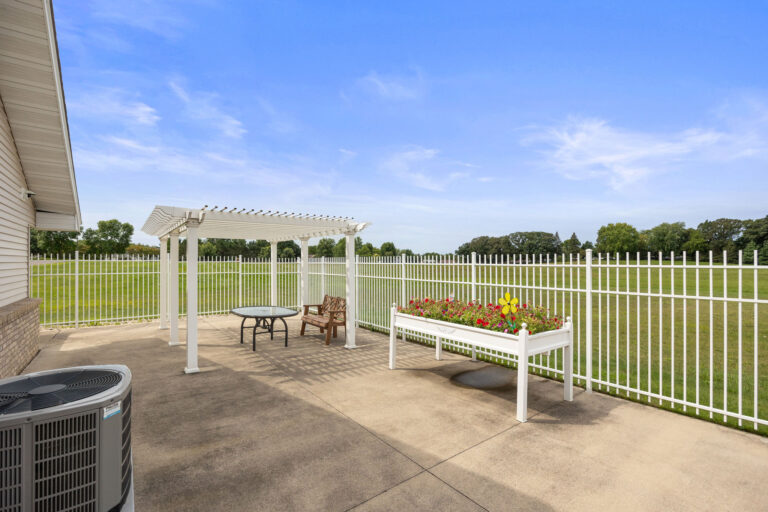 Benedict Homes backyard patio with a white pergola, table, chairs, flower bed, and a white fence overlooking a grassy area.