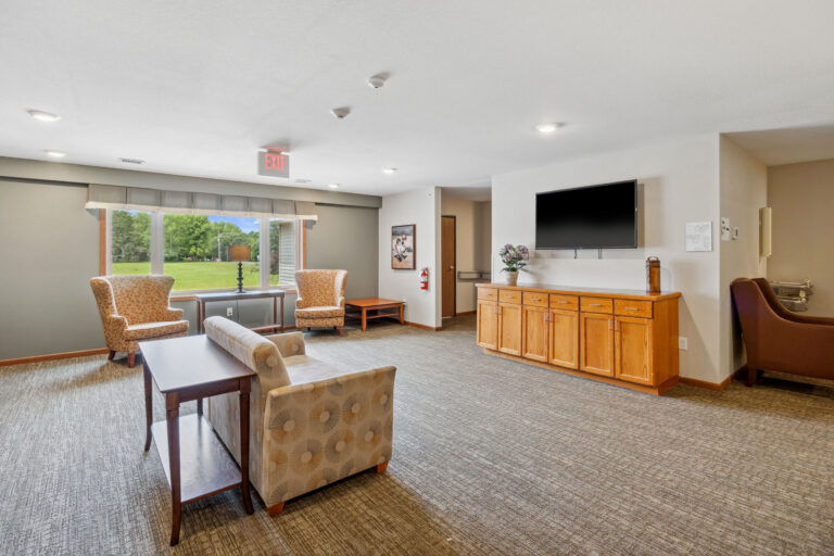 Bright common room with sofas, armchairs, a flat-screen TV, and large window overlooking greenery.