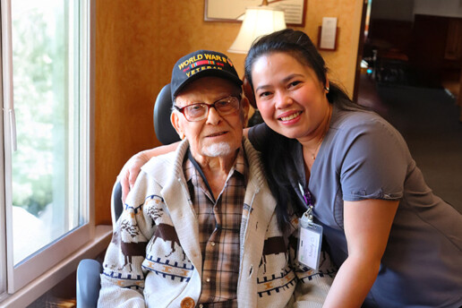A young female Ecumen team member poses for a photo with her arm around an older man with a patch of white beard on his chin, eye glasses and ball cap that says World War II Veteran.