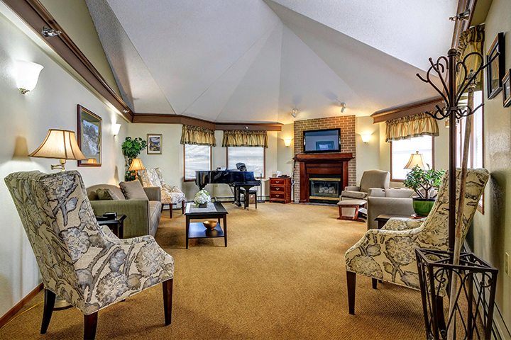 A cozy room with a vaulted ceiling, fireplace, baby grand piano and upholstered lounge chairs.