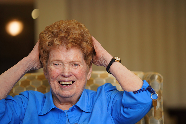 A smiling older woman in a blue blouse, touches her red hair while sitting in an upholstered lounge chair.