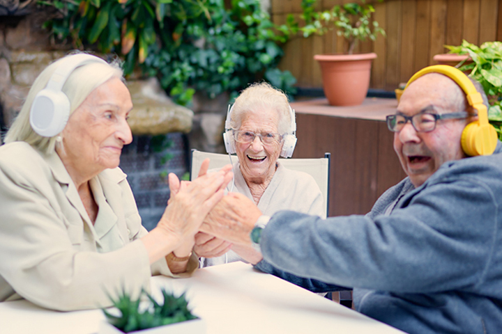 Three older people wearing headphones laugh and clap hands together while sitting at a table outdoors.