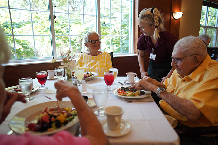 Two older couples dining together, served by a waitress, in a brightly lit room with large windows.