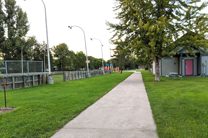 A sidewalk stretches through Westgate Park surrounded by green grass and running between an outdoor hockey rink on the left and a park building on the right.