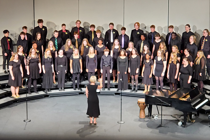 A balcony view of a performing stage where a choir conductor stands next to a grand piano with her back to us, directing high school girls and boys all wearing black standing in four rows of risers as they sing.