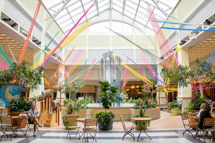 The interior atrium of a shopping mall features a two-story glass ceiling, coloring streamers decorating from floor to ceiling, a central water fountain, large potted plants, and seating areas where people gather to rest and snack.