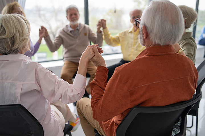 A group of older adults sit in a circle holding hands in prayer, the large windows behind them let in sunlight.