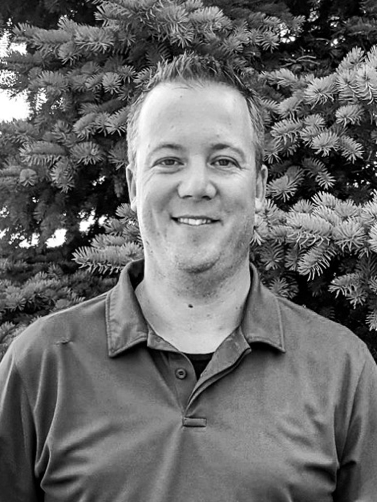 A man with short-cropped and spiked hair, wearing a dark-colored polo-style shirt, smiles for an outdoor portrait in front of a pine tree. Black and white photo.
