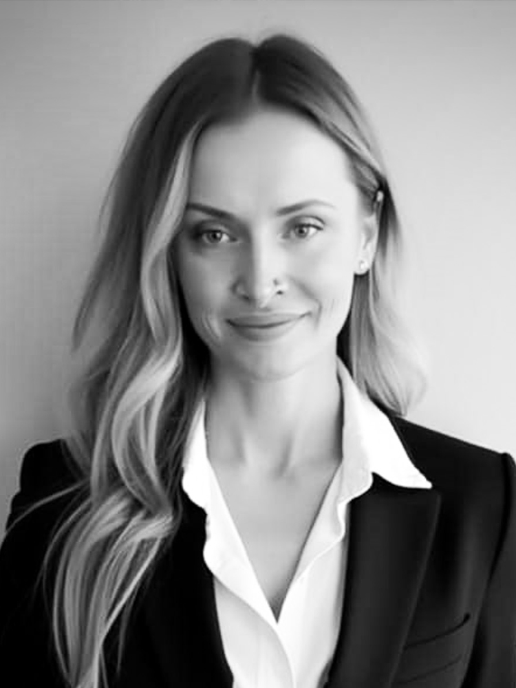 A woman with long blond hair, wearing a white collar shirt and black suit jacket, smiles for an indoor portrait. Black and white.