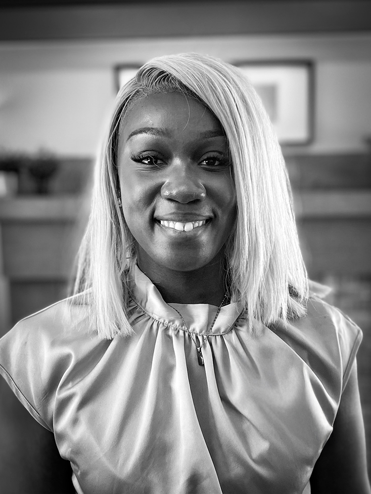 A woman with shoulder-length blond hair and wearing a silk, sleeveless blouse smiles for an indoor portrait. Black and white.