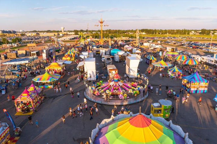 An aerial view of the Red River Valley Fair, featuring brightly colored striped canopies over rides, games and food vendors. Families walk among the structures.