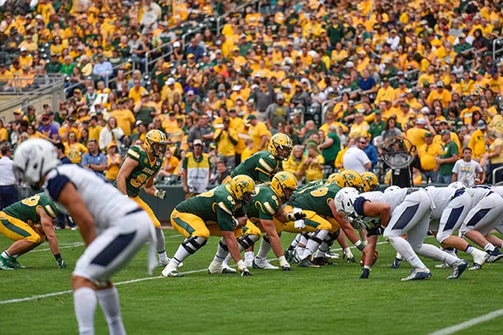 NDSU football offense players in gold helmets, green jerseys and gold pants at the line of scrimmage against a visiting team's defense wearing white helmets, white jerseys and white pants. A sea of gold and green NDSU fans fill the stadium seating in the background.