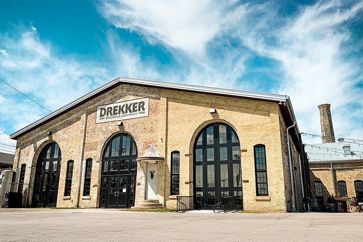 An yellow brick building with three, tall arched windows and a peaked roof with adjoining building featuring a brick smoke stack. A sign painted on the front of the building above the windows reads Drekker Brewing Company.