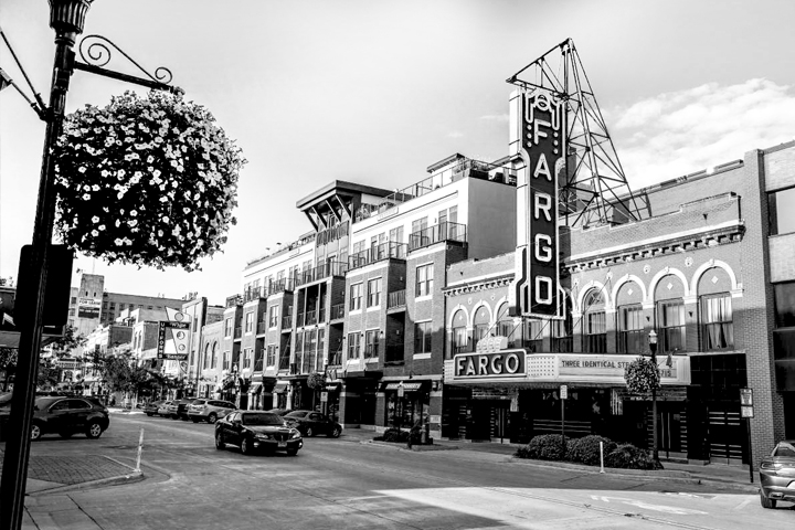A day-time, main street view of the historic Fargo theatre and its neon marquee signs. A care passing by on the road. Parked cars are angled off a street lined with old and new buildings. A flower planter hangs from the light post in the foreground. Black and white photo.