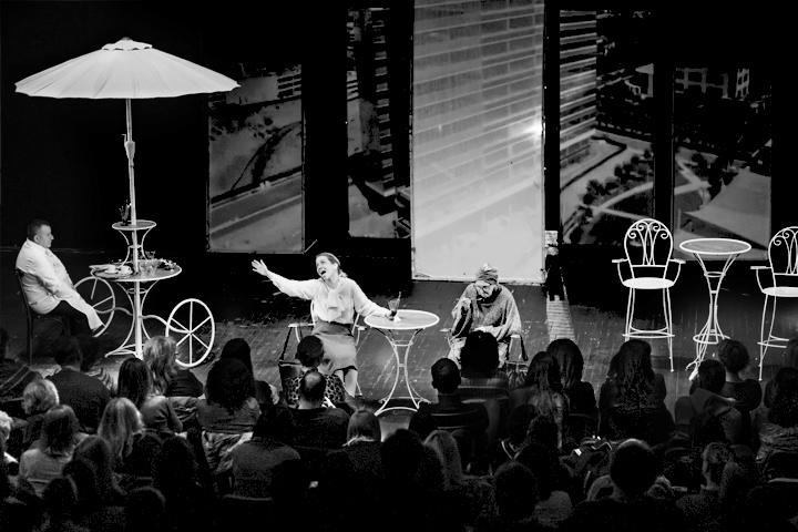 An audience in the foreground watches a young female performer on stage theatrically gesture as she sits at an outdoor cafe with an older female performer watched by a male performer at a neighboring table featuring an umbrella. Black and white photo.